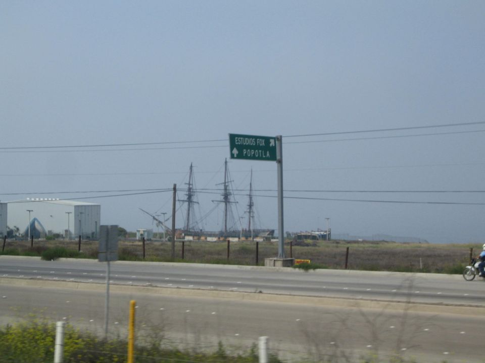 Green highway sign reading ESTUDIOS FOX POPOTLA beside tall ship masts and a white studio building across a field
