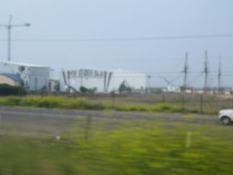 1990 Ford Bronco - motion-blurred roadside view of a large white studio building with a construction crane, seen across a green field with a passing white tru…