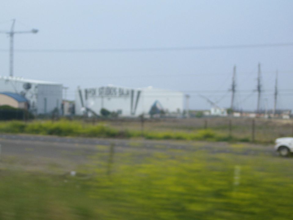 Blurry roadside view of a white film studio building with a crane and a passing white truck across a grassy field