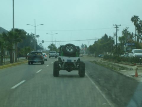 1990 Ford Bronco - Seen through a windshield, a silver tube-chassis rock buggy with a rear-mounted spare tire drives ahead on a palm-lined road in traffic.