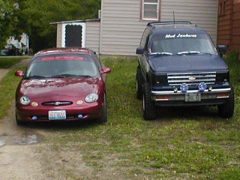 1998 Ford Taurus - Two vehicles, a red Ford Taurus and a blue Chevrolet S-10 Blazer, are parked in front of a building.