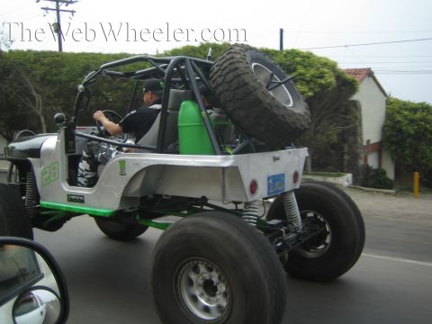 1990 Ford Bronco - driver pilots a silver-and-green tube-chassis rock buggy with oversized tires and a rear-mounted spare down a suburban street.