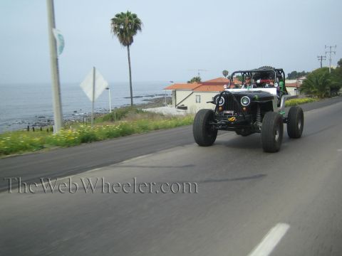 1990 Ford Bronco - silver tube-chassis rock buggy with round headlights speeds along a coastal road lined with a palm tree and the ocean beyond.