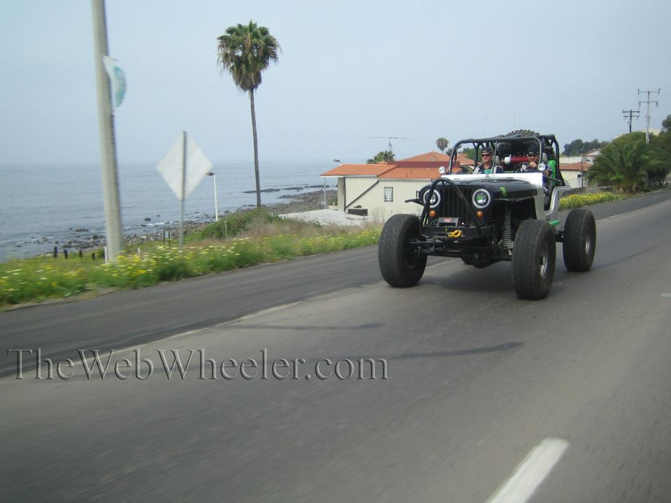 Silver tube-chassis rock buggy with round headlights driving down a coastal road past a palm tree, TheWebWheeler.com watermark