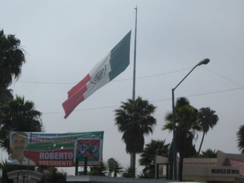 1990 Ford Bronco - large Mexican flag flies atop a tall pole above palm trees, with a ROBERTO PRESIDENTE political campaign banner mounted below.