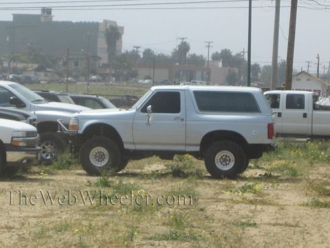 1990 Ford Bronco - silver lifted Ford Bronco with a removable hardtop sits parked on a dirt lot among pickups, with a town and palms in the background.