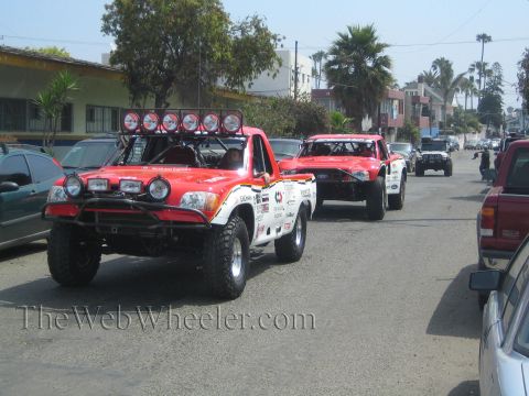1990 Ford Bronco - Two red off-road prerunner race trucks with roof light bars cruise down a street lined with parked cars and palm trees.