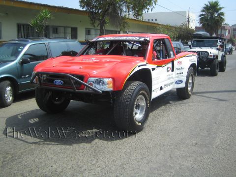 1990 Ford Bronco - red Ford prerunner race truck covered in sponsor decals, with a roof light bar, sits on a street next to a white rock buggy.