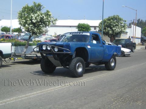 1990 Ford Bronco - blue lifted prerunner truck with a tubular front bumper and round lights sits on a street beside a flatbed trailer and parked cars.
