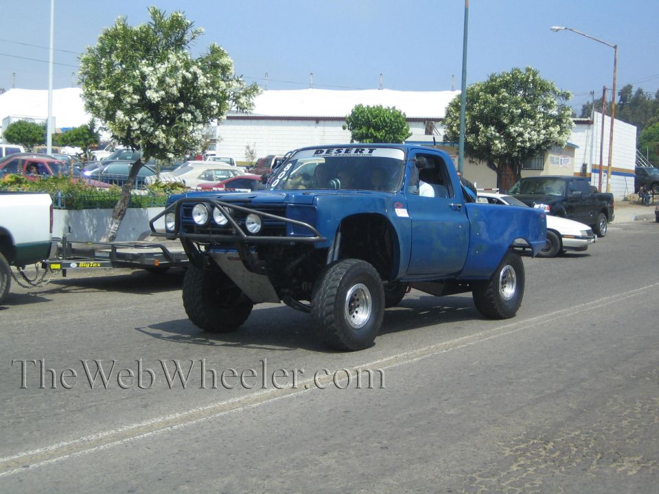 Blue lifted prerunner truck with tube front bumper and round lights parked on a street near a flatbed trailer