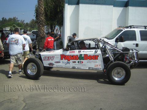 1990 Ford Bronco - white single-seat tube-chassis off-road race buggy plastered with Papas and Beer and BFGoodrich decals sits on a street as people stand aro…