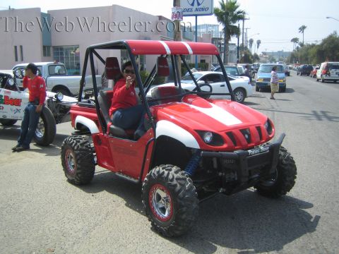 1990 Ford Bronco - red side-by-side UTV with a white hood stripe and a seated rider sits on a busy street with shops, palms and parked cars behind.