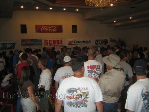 1990 Ford Bronco - crowd gathers indoors beneath a chandelier at an off-road racing event, with SCORE, BFGoodrich and Coca-Cola banners on the wall.