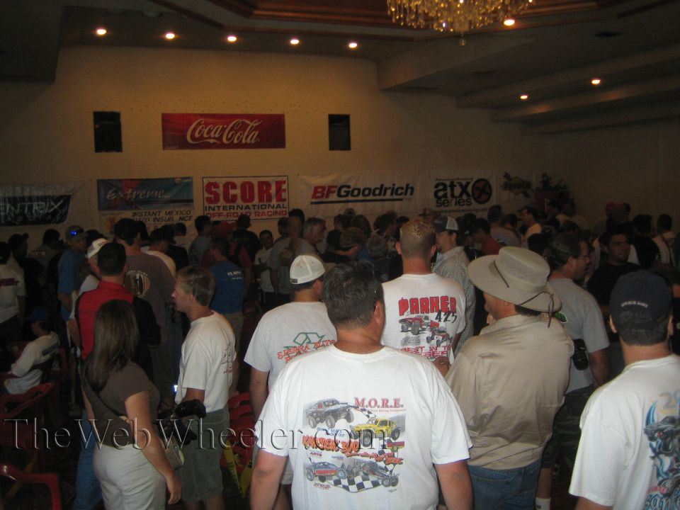 Crowd of people indoors at an event under a chandelier, with SCORE, BFGoodrich and Coca-Cola banners on the wall