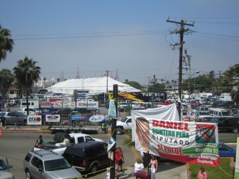 1990 Ford Bronco - outdoor event lot crowded with vehicles, a large white tent, SCORE and BFGoodrich banners, palm trees and a political campaign banner.