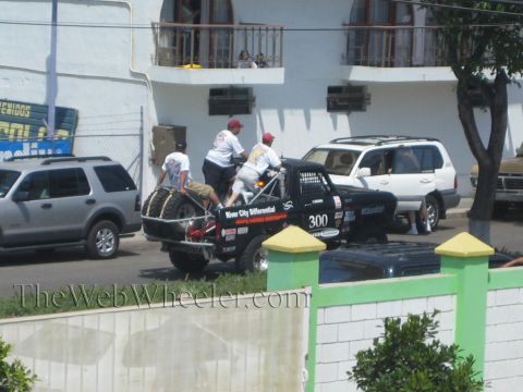 1990 Ford Bronco - Crew members tend to a black number 300 off-road race truck loaded on a trailer outside a building, with an SUV parked nearby.