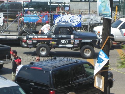 1990 Ford Bronco - black number 300 off-road race truck marked River City Differential and BFGoodrich sits in a fenced staging lot.
