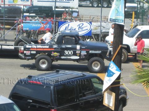 1990 Ford Bronco - black number 300 race truck with sponsor decals waits near a fence as a black SUV and dirt bikes sit in the background.