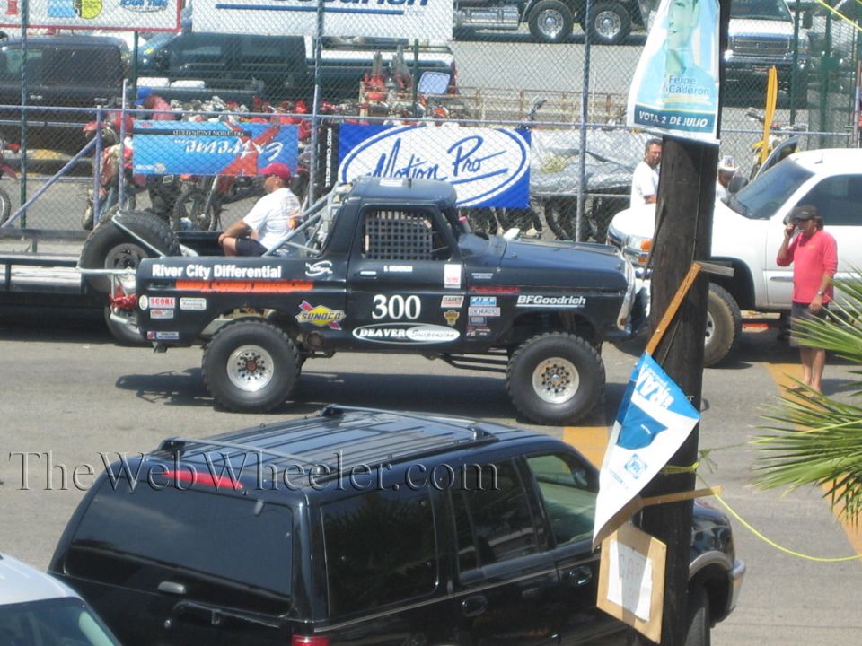 Black #300 off-road race truck in a fenced staging area with dirt bikes behind