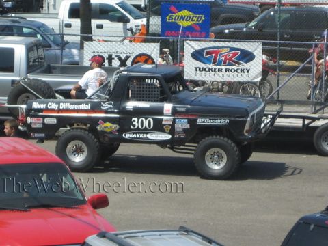 1990 Ford Bronco - black number 300 off-road race truck with River City Differential decals sits in a paddock near Sunoco and Tucker Rocky banners.