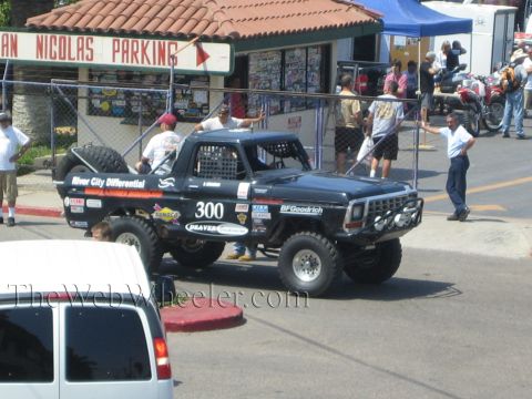 1990 Ford Bronco - black number 300 lifted off-road race truck idles on a street as bystanders watch, beneath a San Nicolas Parking sign.