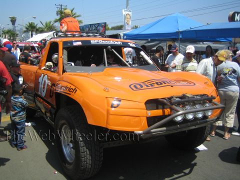 1990 Ford Bronco - orange number 10 off-road trophy truck with a roof light bar and tube bumper draws a crowd at an outdoor event.