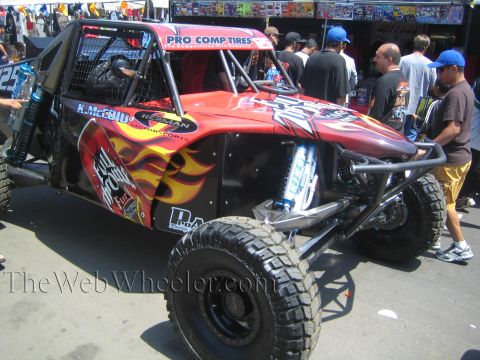 1990 Ford Bronco - red and black off-road race buggy with flame graphics and Pro Comp tires sits among onlookers at an outdoor venue.