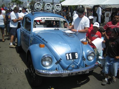 1990 Ford Bronco - light blue Baja-prepped VW Beetle number 119 with Hella lights draws a crowd as a man signs paperwork on the hood.