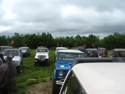 1992 Ford Bronco - A collection of off-road vehicles, including a blue 1992 Ford Bronco, gathered in a grassy area under a cloudy sky.