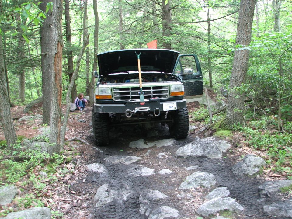 Ford F-Series truck with hood up on a rocky, wooded trail. A shovel is in the engine bay