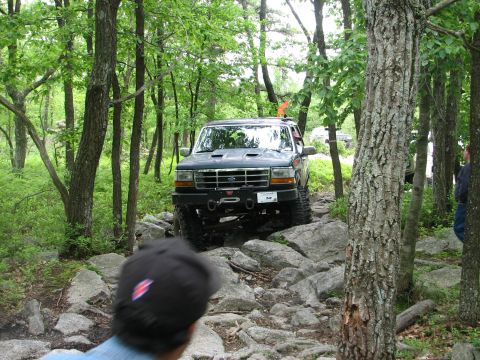 1992 Ford Bronco - A 1992 Ford Bronco tackles a challenging rocky path through a dense forest, with another vehicle visible in the background.