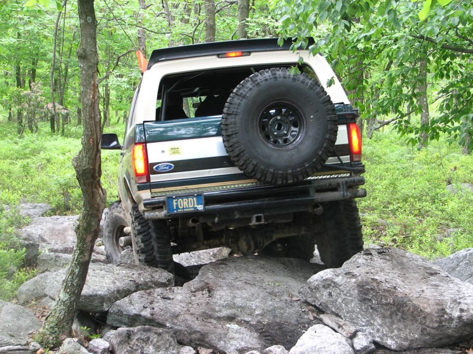 Rear of a lifted green and tan Ford Bronco with swing-out spare crawling over rocks in the woods