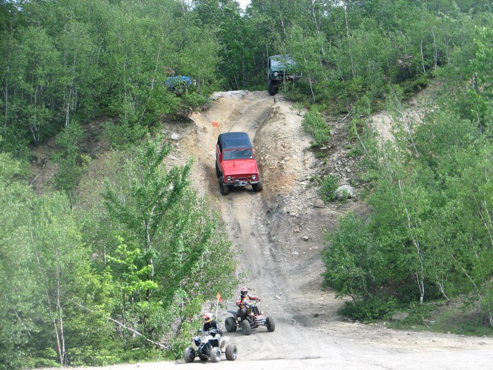 Red soft-top Jeep with winch descending a steep dirt hillclimb as two ATVs wait below