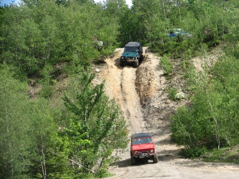 1992 Ford Bronco - Multiple off-road vehicles navigate a steep, rutted dirt path surrounded by lush green trees.