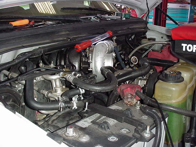Engine bay with various hoses, battery, and tools on top of the engine cover. Text 'SUPERMOTORS' visible