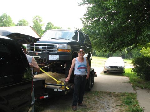1992 Ford Bronco - A dark green 1992 Ford Bronco is loaded onto a trailer, with a man standing beside it and another vehicle parked in the background.