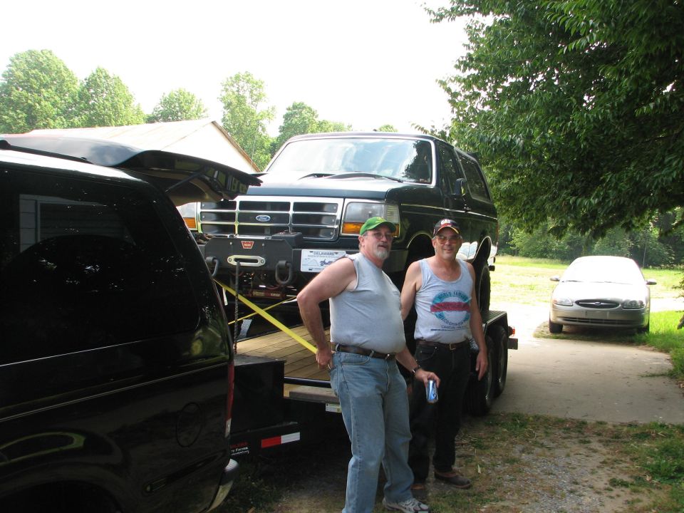Dark green Ford Bronco with winch bumper strapped to a trailer behind a black tow SUV