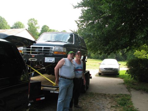 1992 Ford Bronco - A dark green Ford Bronco is loaded onto a trailer, with two men standing nearby and a silver sedan in the background.