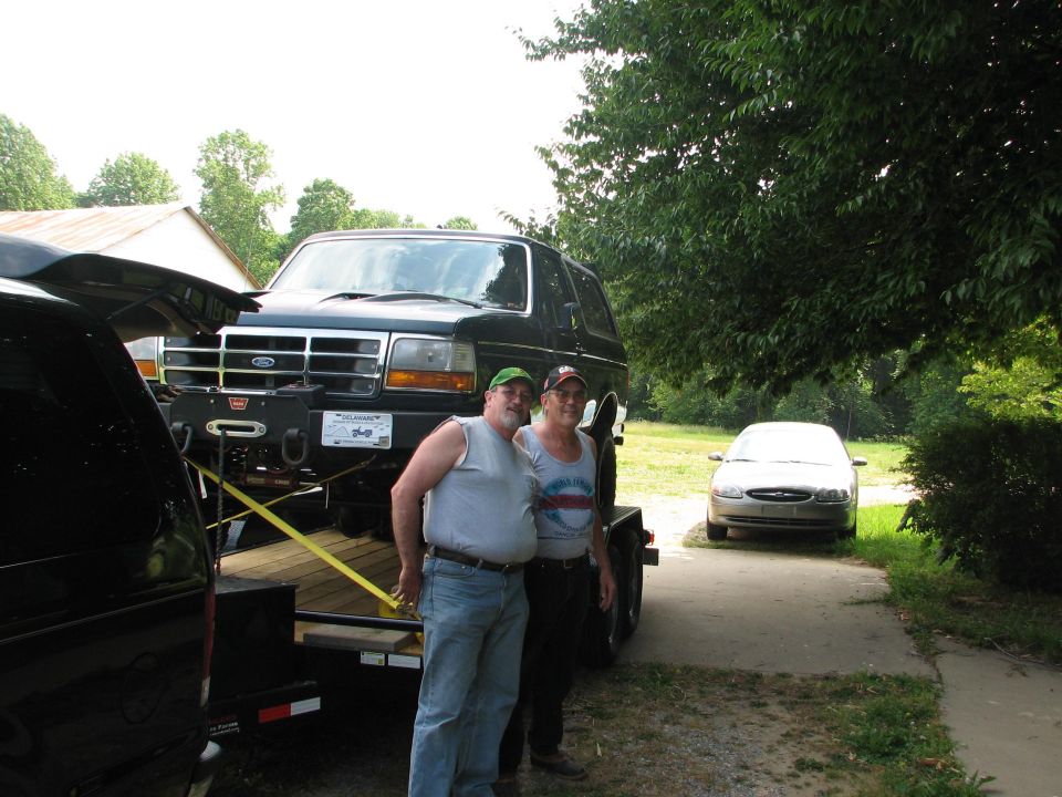 Two men in front of a dark Ford Bronco with winch bumper loaded on a trailer in a gravel drive