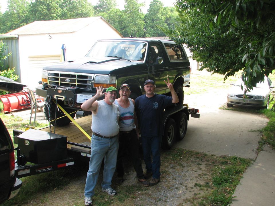 Dark green Ford Bronco with winch bumper strapped on a tandem-axle trailer, three men posing