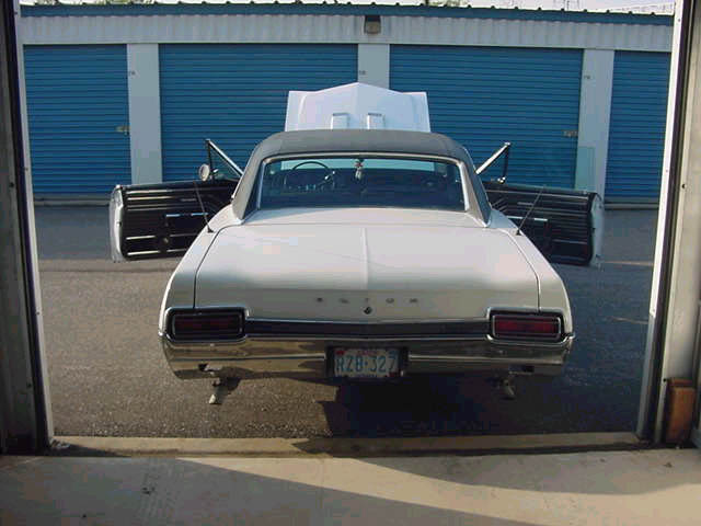 White coupe with black roof, open doors, and hood up. Rear view shows chrome bumper and license plate RZB-327