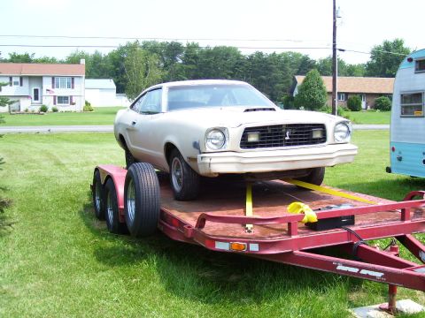 1978 Ford Mustang II - A beige coupe with a hood scoop is loaded onto a red trailer in a grassy area.