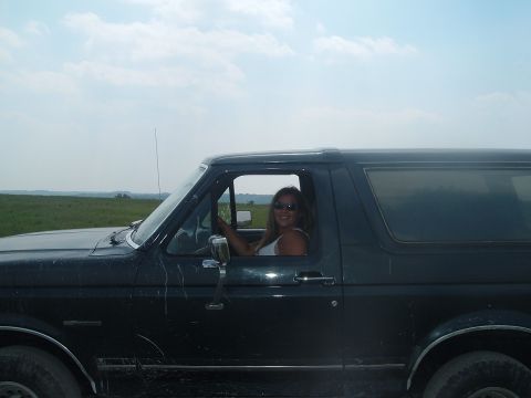 1988 Ford Bronco - A woman smiles while driving a dark 1988 Ford Bronco through a grassy field under a cloudy sky.