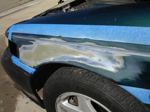 1995 Ford Taurus - The fender of a dark green car is taped off and sanded down, showing metal and primer before repair.