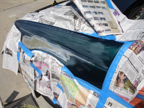 1995 Ford Taurus - A car fender is masked with newspaper and blue tape, revealing areas of primer and fresh dark green paint during a repair process.