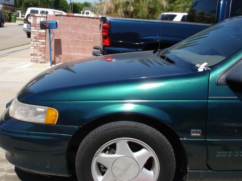 1995 Ford Taurus - A close-up view of a green sedan's fender and hood, with other vehicles parked behind it.