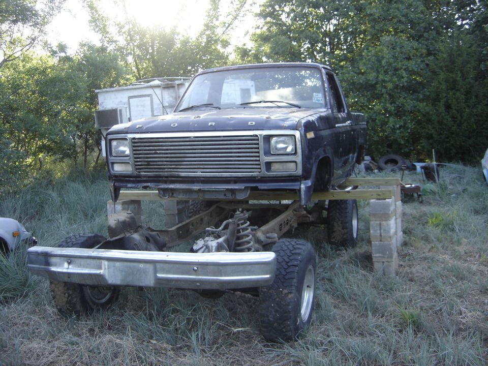 Dark blue Ford truck body on wood beams and cinder blocks above a separated chassis and axle in a grassy yard