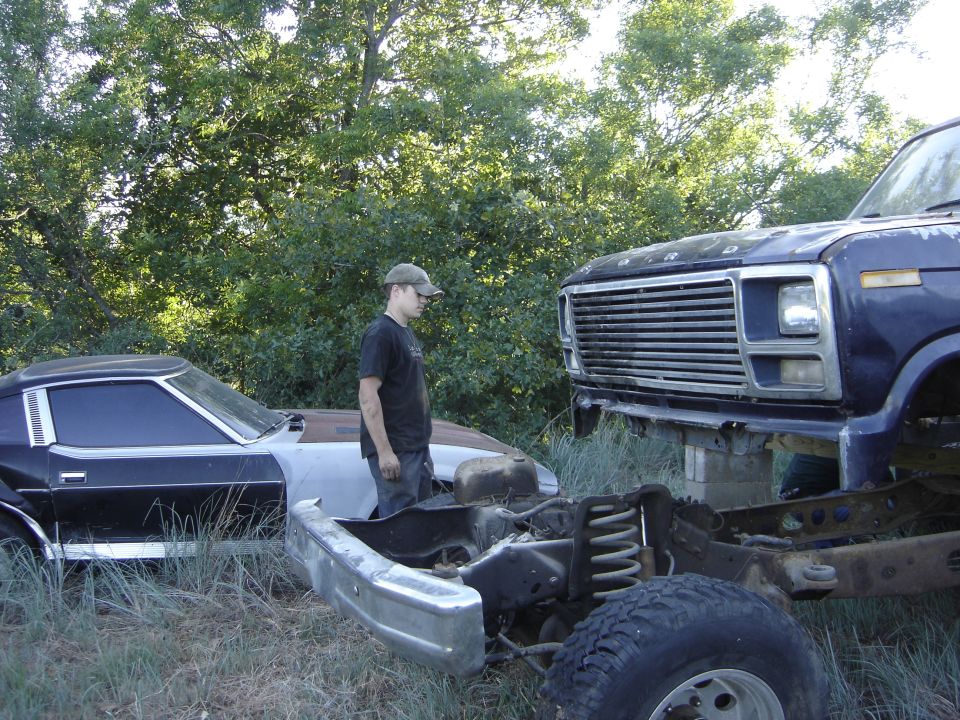 Man inspecting a derelict van/truck cab and a stripped sedan in an overgrown lot