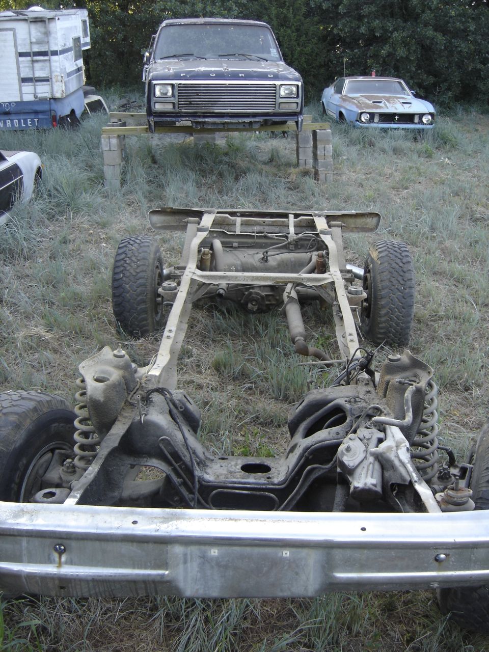 Bare Ford rolling chassis in grass; truck body on blocks and blue Mustang in background