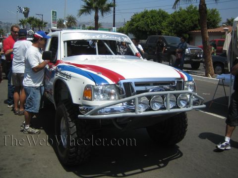 1990 Ford Bronco - white Ford Ranger prerunner with a red, white and blue stripe and front light pods is prepped on a street by its crew.